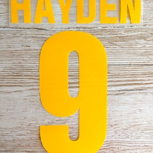 Może przedstawiać: Żółte litery termotransferowe tworzące napis "HAYDEN" i cyfrę "9" na drewnianym tle.
