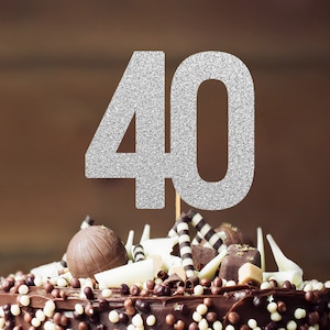 May include: A chocolate cake decorated with chocolate sprinkles, chocolate curls, and chocolate candies. A silver glitter cake topper with the number "40" is on top of the cake.