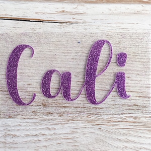 May include: Purple glitter vinyl decal with the word "Cali" in a cursive font.