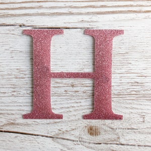 May include: A pink glitter letter H on a white wood background.