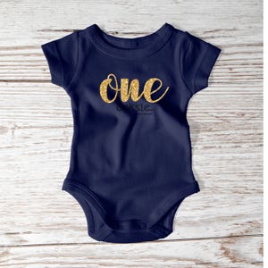 May include: Navy blue baby bodysuit with the word "ONE" in gold glitter script. The bodysuit has short sleeves and a snap closure. The word "oksie" is printed below "ONE".