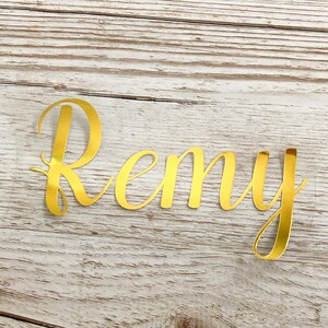 May include: Gold foil letters spelling the name "Remy" on a light wood background.