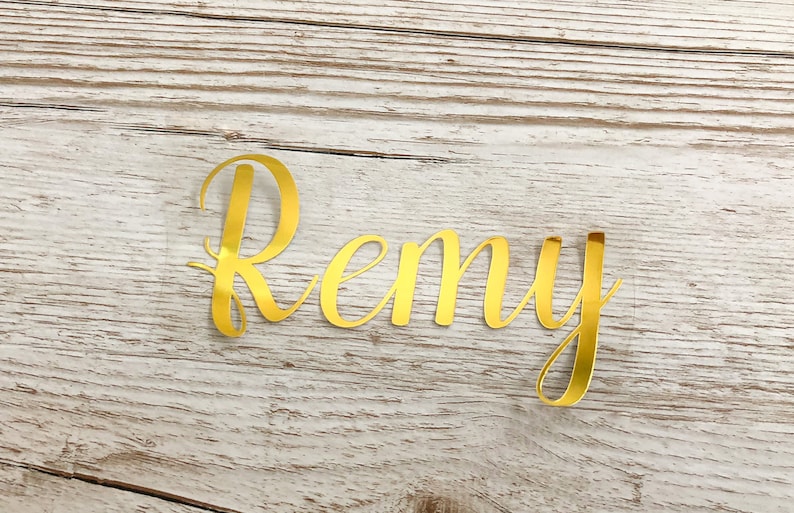 May include: Gold foil letters spelling the name "Remy" on a light wood background.