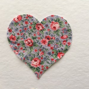 以下が含まれることがあります： 花柄のハート型の布のパッチ。布は薄い青色で、小さな白いドットがあり、赤いバラとピンクのバラに緑の葉が描かれています。
