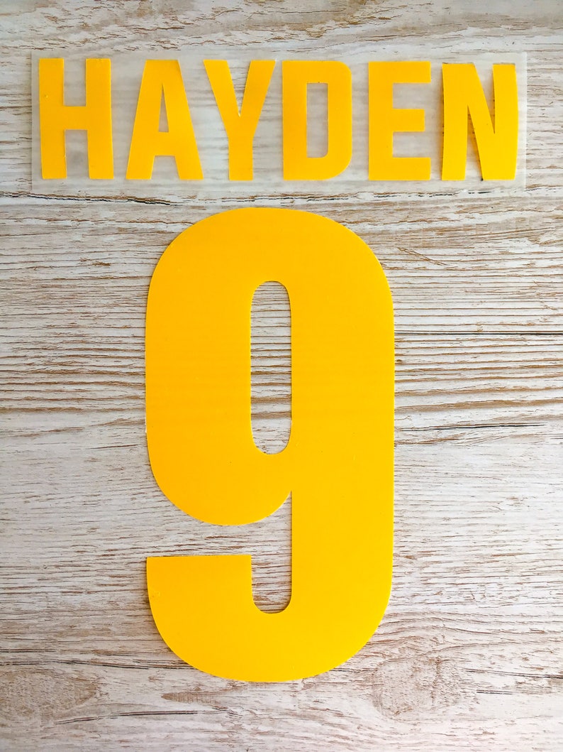 May include: Yellow iron-on letters spelling "HAYDEN" and the number "9" on a wood background.