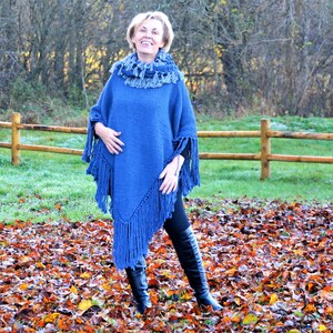 May include: A blue knit poncho with fringe detailing. The poncho has a cowl neck and is paired with a matching scarf. The model is wearing black pants and black boots. The background features a wooden fence and autumn leaves.