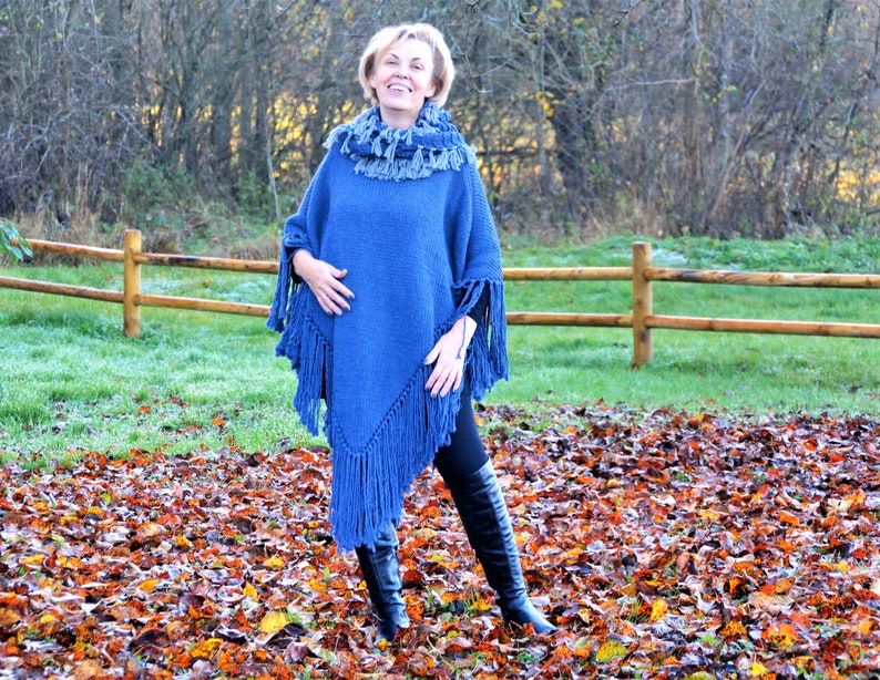 May include: A blue knit poncho with fringe detailing. The poncho has a cowl neck and is paired with a matching scarf. The model is wearing black pants and black boots. The background features a wooden fence and autumn leaves.