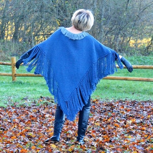 May include: A blue knitted poncho with fringe detail. The poncho has a round neckline and is worn with black gloves and boots. The background features a wooden fence, green grass, and fallen leaves.