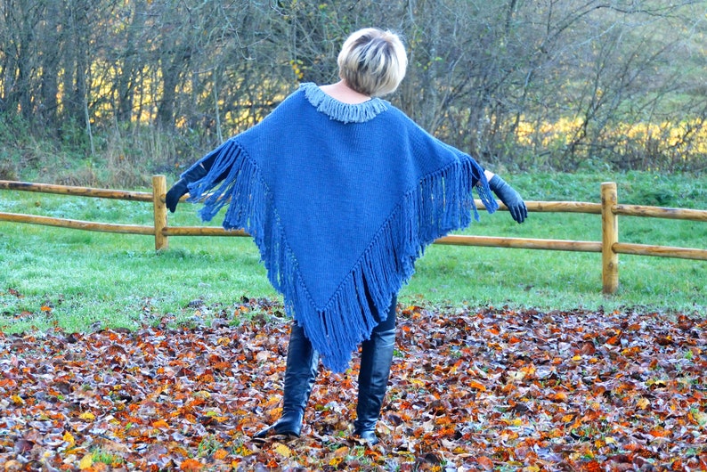 May include: A blue knitted poncho with fringe detail. The poncho has a round neckline and is worn with black gloves and boots. The background features a wooden fence, green grass, and fallen leaves.