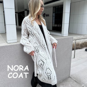 May include: A white open knit cardigan with a long, flowing silhouette. The cardigan has a V-neck and a tie belt. The text "Nora Coat" is displayed on the image.