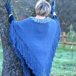 May include: A blue knitted poncho with fringe detailing. The poncho has a fringed collar and a triangular shape, with long fringe along the edges. The garment is a solid blue color and appears to be made of a soft, warm material.