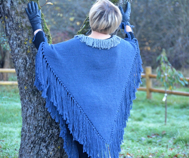 May include: A blue knitted poncho with fringe detailing. The poncho has a fringed collar and a triangular shape, with long fringe along the edges. The garment is a solid blue color and appears to be made of a soft, warm material.