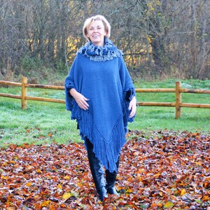 May include: A blue knitted poncho with fringe detailing. The poncho is worn with a matching scarf and black boots. The background features a wooden fence, grass, and autumn leaves.