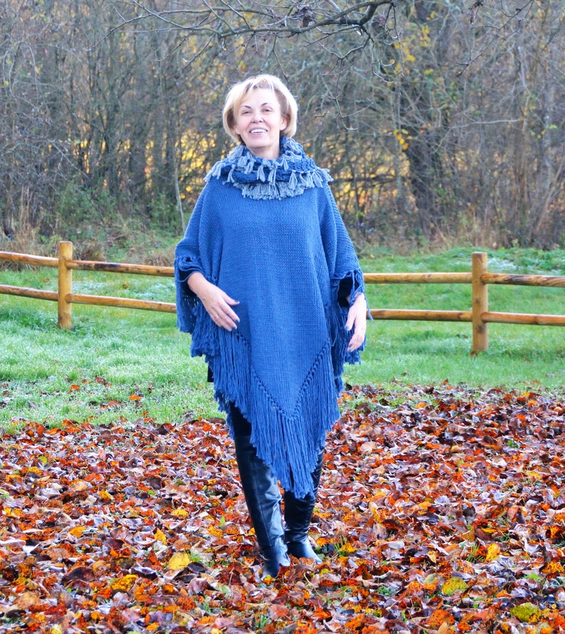 May include: A blue knitted poncho with fringe detailing. The poncho is worn with a matching scarf and black boots. The background features a wooden fence, grass, and autumn leaves.