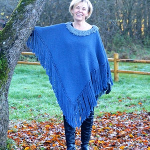 May include: A blue knitted poncho with fringe detailing. The poncho has a round neckline with fringe and is worn with black boots. The background includes a tree, grass, and fallen leaves.