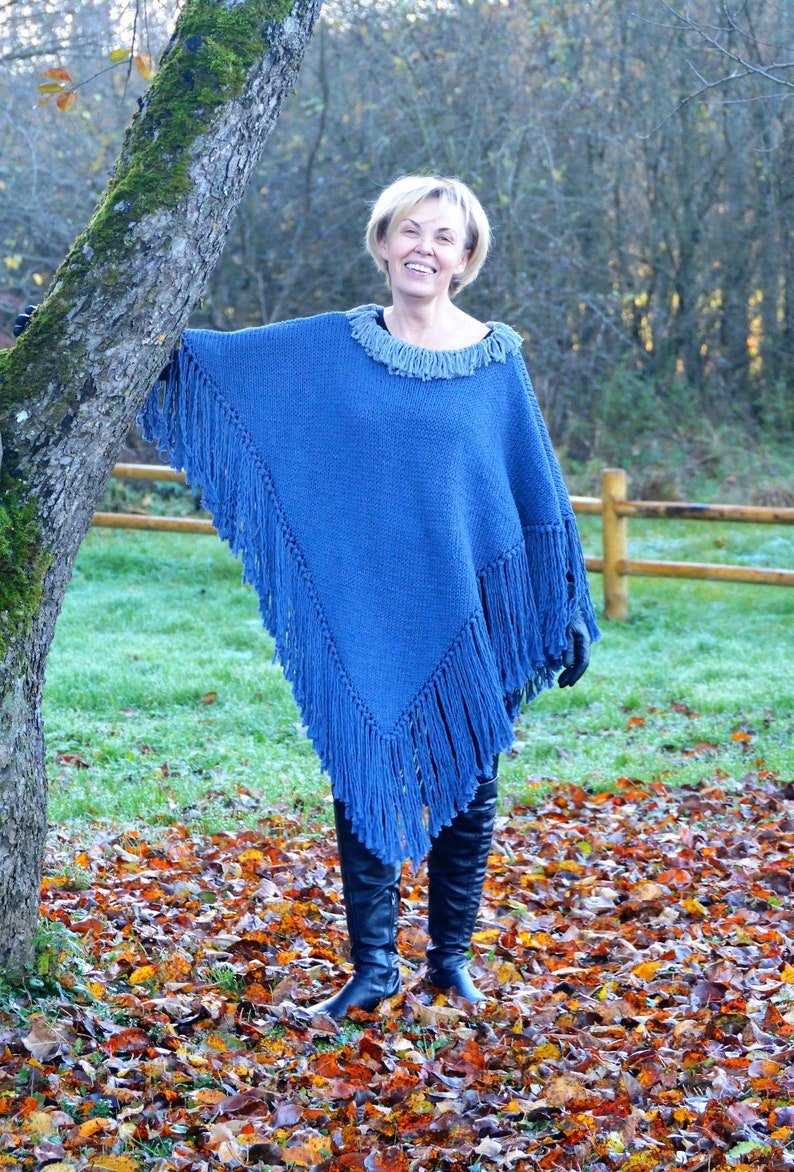 May include: A blue knitted poncho with fringe detailing. The poncho has a round neckline with fringe and is worn with black boots. The background includes a tree, grass, and fallen leaves.