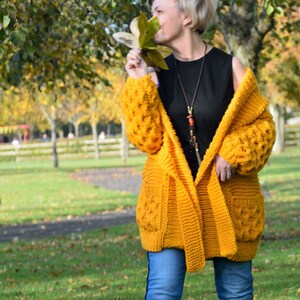May include: A woman wearing a yellow knitted cardigan with a black tank top and blue jeans. She is holding a leaf in her hand and smiling.