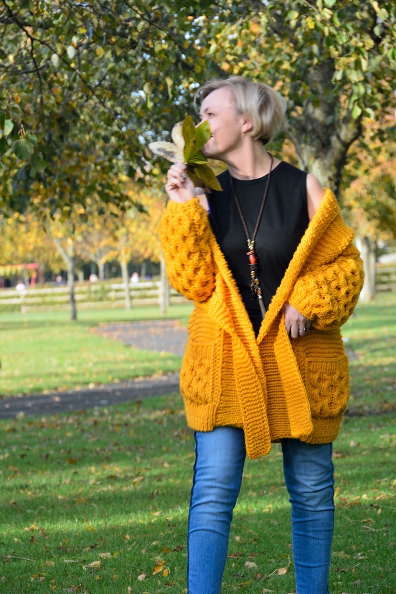 May include: A woman wearing a yellow knitted cardigan with a black tank top and blue jeans. She is holding a leaf in her hand and smiling.