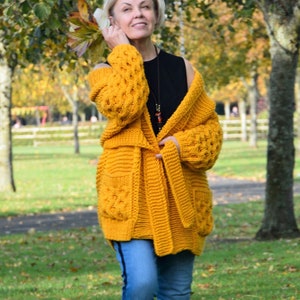 May include: A woman wearing a yellow knitted cardigan with a black top and blue jeans. The cardigan has a cable knit pattern and a tie belt.