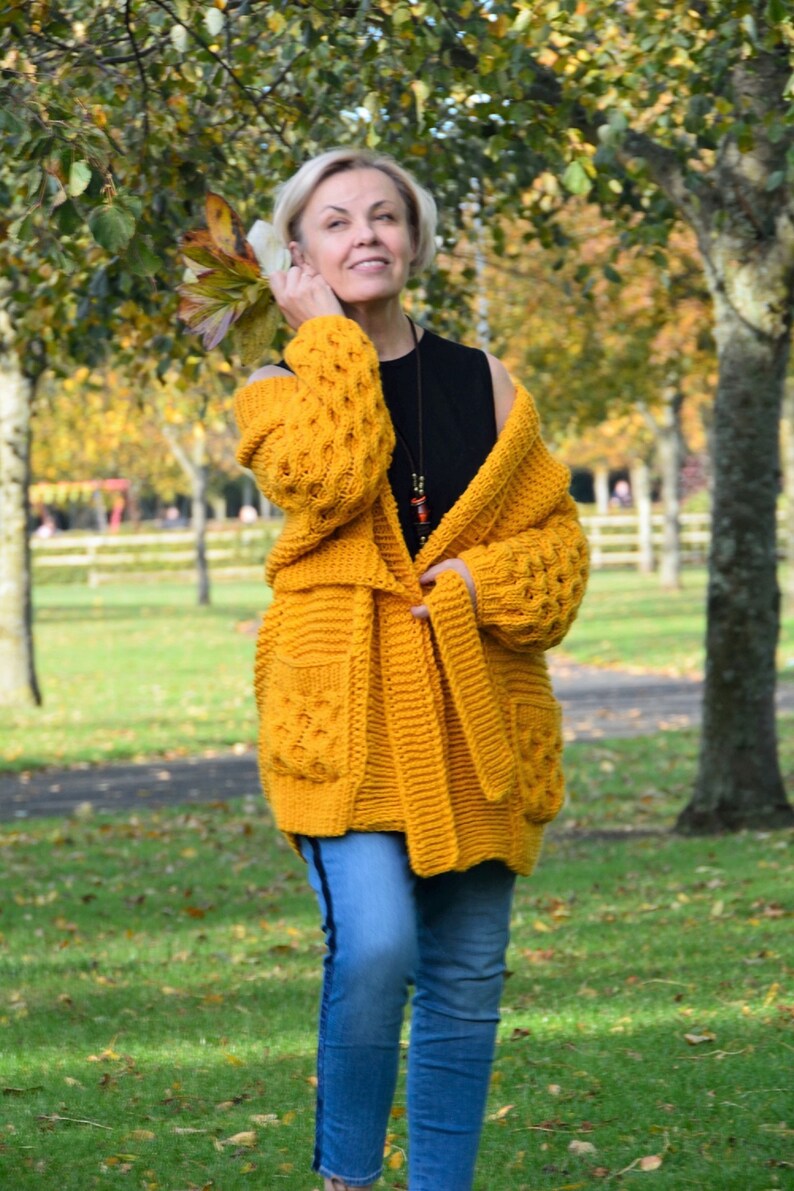May include: A woman wearing a yellow knitted cardigan with a black top and blue jeans. The cardigan has a cable knit pattern and a tie belt.