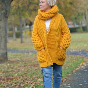 May include: A woman wearing a mustard yellow cardigan with a matching scarf and blue jeans. The cardigan has a shawl collar and pockets. The woman is walking on a path in a park.