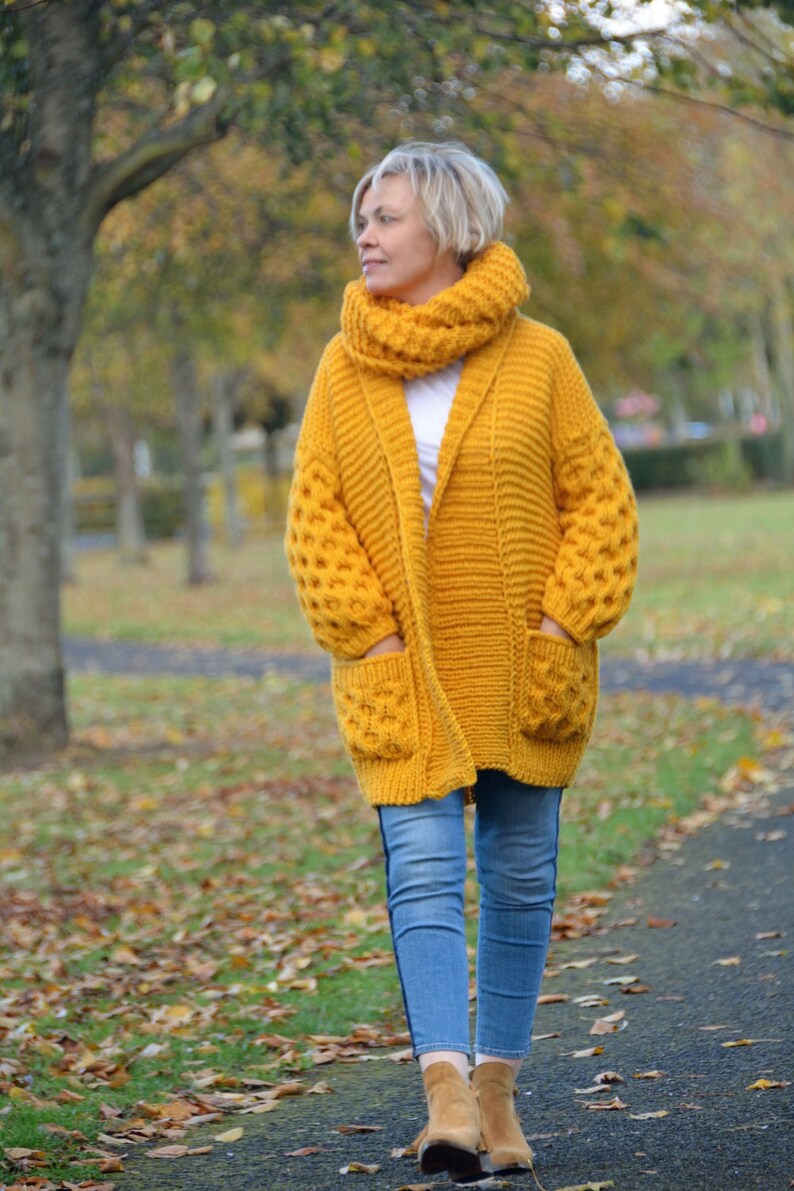 May include: A woman wearing a mustard yellow cardigan with a matching scarf and blue jeans. The cardigan has a shawl collar and pockets. The woman is walking on a path in a park.