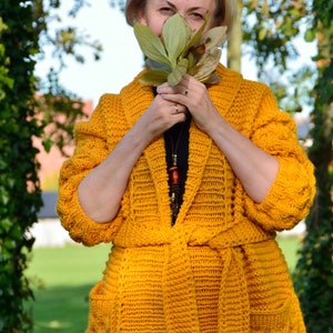 May include: A mustard yellow crocheted cardigan with a tie belt and pockets. The cardigan is made with a chunky yarn and has a relaxed fit.
