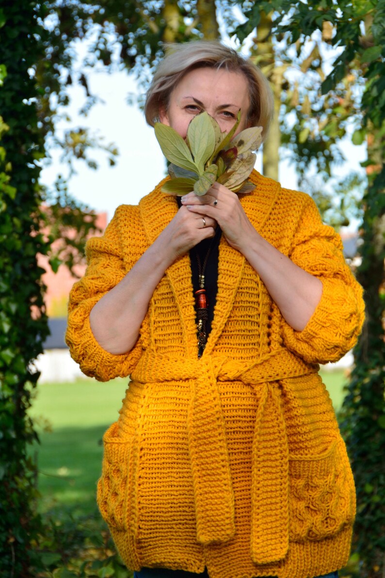 May include: A mustard yellow crocheted cardigan with a tie belt and pockets. The cardigan is made with a chunky yarn and has a relaxed fit.