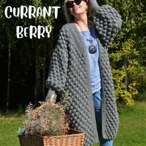 May include: A gray knitted cardigan with a textured bubble stitch pattern. The cardigan has a long, open front and long sleeves. The model is wearing the cardigan over a blue shirt and jeans.