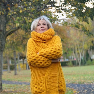 May include: A long, yellow knitted cardigan with a large, chunky knit pattern. The cardigan has a high collar and a loose fit. The cardigan is worn by a person standing in a park with trees in the background.