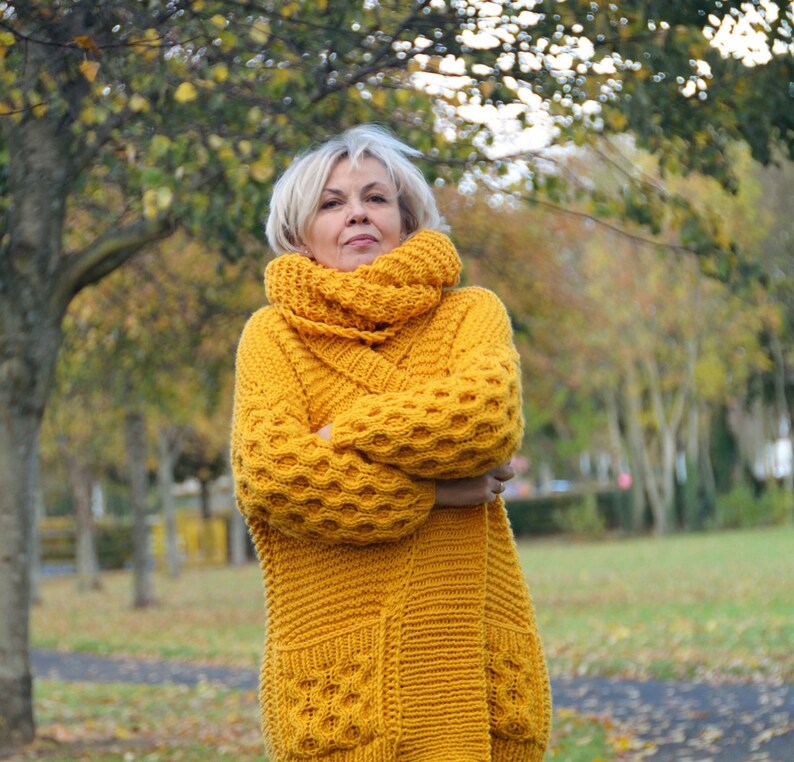 May include: A long, yellow knitted cardigan with a large, chunky knit pattern. The cardigan has a high collar and a loose fit. The cardigan is worn by a person standing in a park with trees in the background.