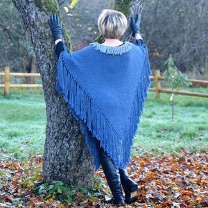 May include: A blue knitted poncho with fringe detail. The poncho has a gray collar and is worn with black gloves and boots. The person is leaning against a tree in an outdoor setting with fall foliage.