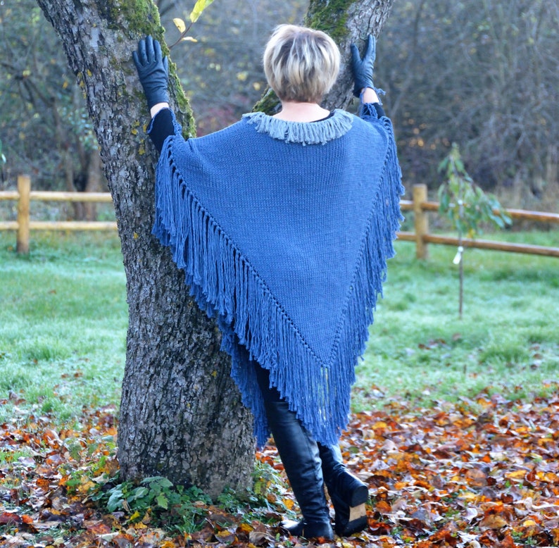 May include: A blue knitted poncho with fringe detail. The poncho has a gray collar and is worn with black gloves and boots. The person is leaning against a tree in an outdoor setting with fall foliage.