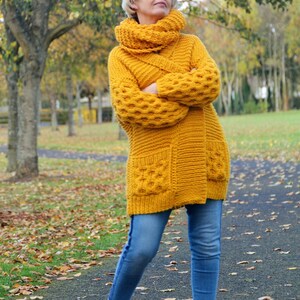 May include: A woman wearing a yellow knitted cardigan with a cowl neck and pockets. She is wearing blue jeans and brown ankle boots. The cardigan has a textured pattern and is a long style.