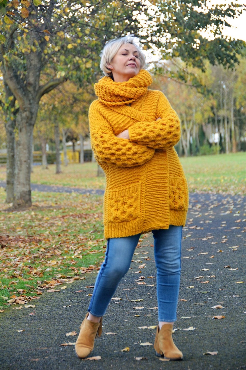 May include: A woman wearing a yellow knitted cardigan with a cowl neck and pockets. She is wearing blue jeans and brown ankle boots. The cardigan has a textured pattern and is a long style.