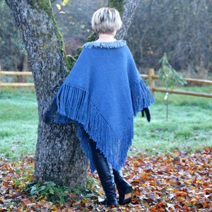 May include: A blue knitted poncho with fringe detailing. The poncho has a fringed collar and fringed edges along the bottom and sleeves. The person is wearing black gloves and black boots. The background includes a tree, a wooden fence, and fallen leaves.
