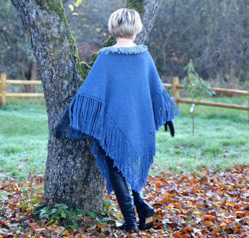 May include: A blue knitted poncho with fringe detailing. The poncho has a fringed collar and fringed edges along the bottom and sleeves. The person is wearing black gloves and black boots. The background includes a tree, a wooden fence, and fallen leaves.