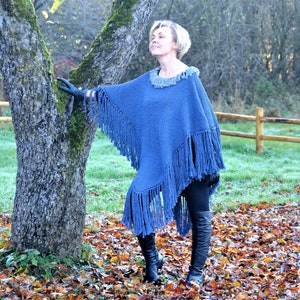 May include: A blue knitted poncho with long fringe detailing. The poncho has a round neckline with a lighter blue trim. The garment is worn outdoors with black leggings and boots. The background includes a tree and fall foliage.