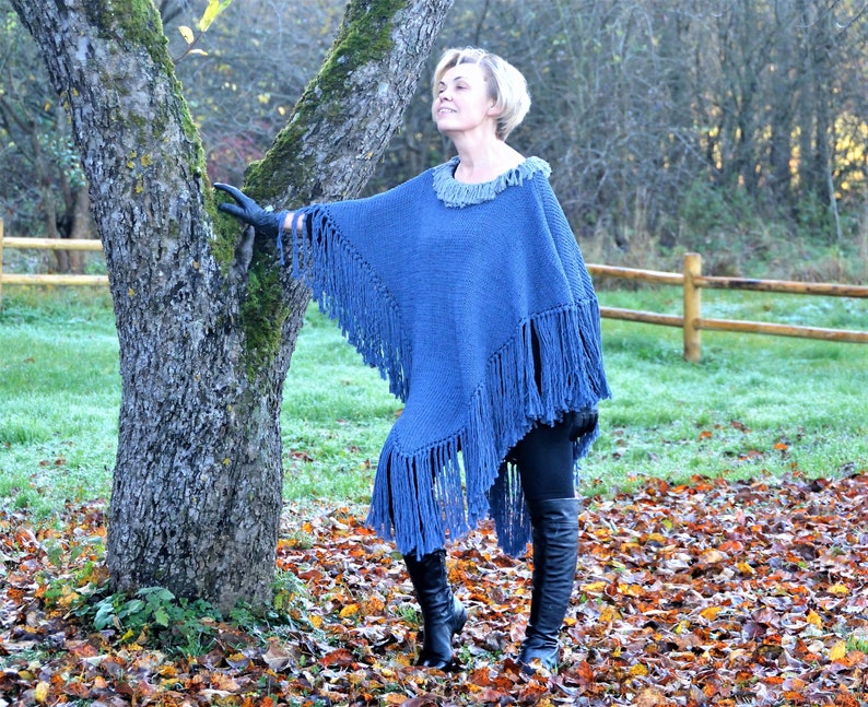 May include: A blue knitted poncho with long fringe detailing. The poncho has a round neckline with a lighter blue trim. The garment is worn outdoors with black leggings and boots. The background includes a tree and fall foliage.