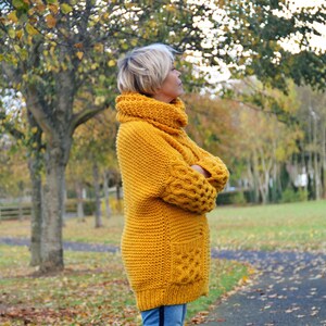 May include: A mustard yellow knitted sweater with a high cowl neck and a large pocket on the front. The sweater has a chunky knit texture and is worn by a person standing in a park.