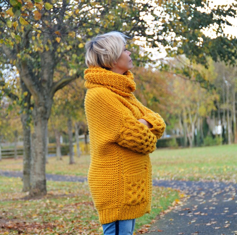 May include: A mustard yellow knitted sweater with a high cowl neck and a large pocket on the front. The sweater has a chunky knit texture and is worn by a person standing in a park.