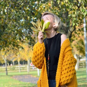 May include: A woman wearing a yellow knitted cardigan with a black tank top and blue jeans. She is holding a bunch of yellow leaves in front of her face.
