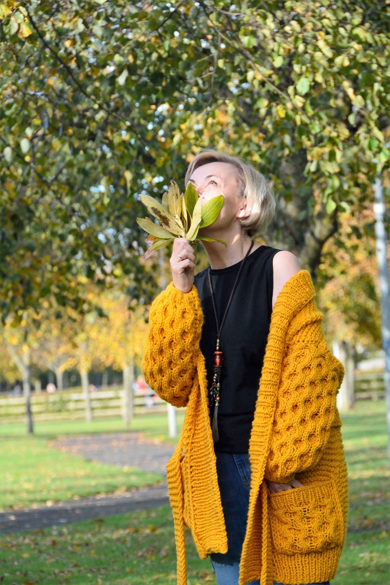 May include: A woman wearing a yellow knitted cardigan with a black tank top and blue jeans. She is holding a bunch of yellow leaves in front of her face.