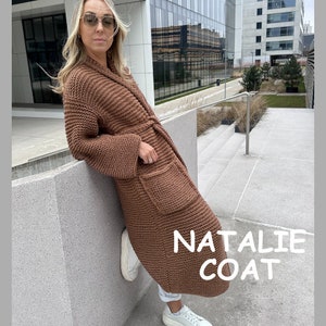 Può includere: Un cappotto marrone a maglia con collo a scialle, maniche lunghe e due tasche anteriori. Il cappotto è lungo fino al ginocchio e ha una cintura abbinata. Il testo "NATALIE COAT" è visualizzato in bianco in basso a destra del cappotto.