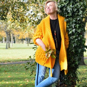 May include: A woman wearing a yellow knitted cardigan with a black necklace and blue jeans stands next to a tree with green ivy. She is holding a bunch of autumn leaves in her hand.