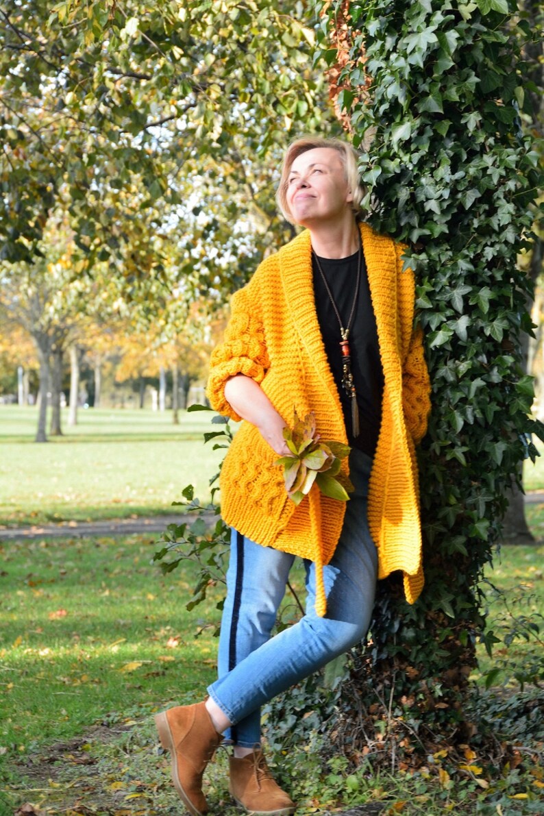 May include: A woman wearing a yellow knitted cardigan with a black necklace and blue jeans stands next to a tree with green ivy. She is holding a bunch of autumn leaves in her hand.