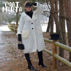 May include: A white, textured coat with large pockets and black buttons. The coat has a wide collar and is worn with black gloves and boots. The text "COAT MARTA" is visible in the upper left corner. A black hat rests on a wooden fence.
