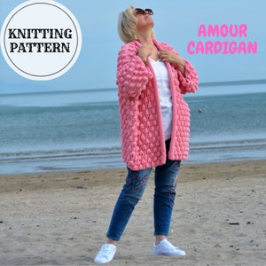 May include: A pink knitted cardigan with a textured bobble stitch pattern. The cardigan is open front and has long sleeves. The model is wearing the cardigan with a white t-shirt and blue jeans. The text "Amour Cardigan" is visible in the image.