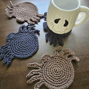 Puede incluir: Una taza de cerámica blanca con un diseño de cara de fantasma descansa sobre un posavasos de ganchillo gris oscuro. También son visibles tres posavasos hechos a mano adicionales en tonos marrones y azules. Los posavasos tienen un diseño único en forma de araña.