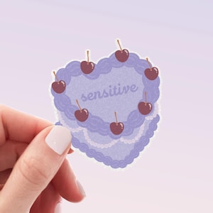May include: Purple heart-shaped cake with white frosting and red cherries. The cake is decorated with the word "sensitive" in white.